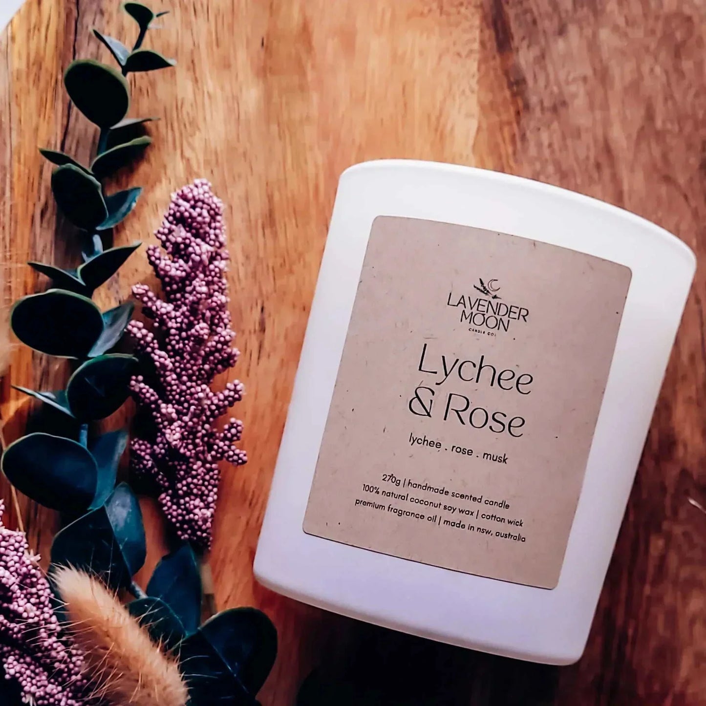 Lychee & Rose Candle with floral and musk scent, vegan-friendly coconut soy wax, on wooden surface with pink and green foliage.