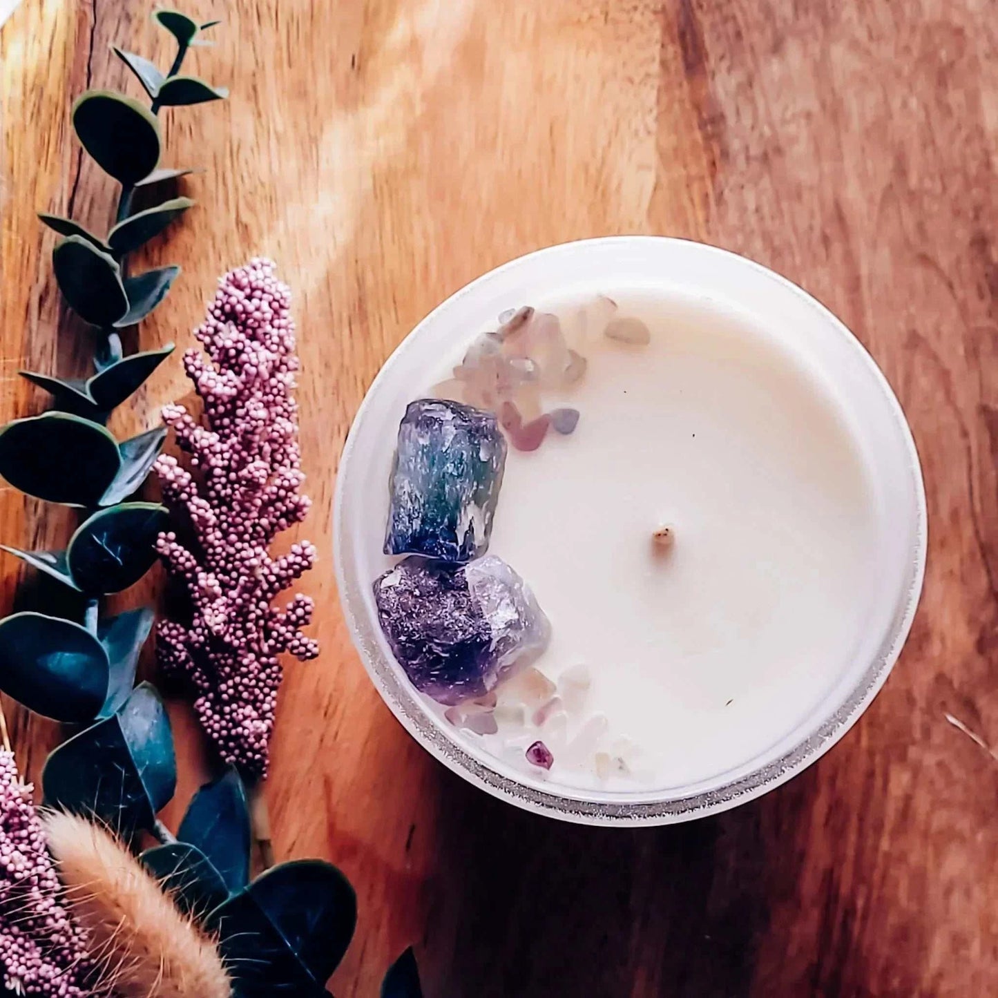 Manifest Candle with Rainbow Flourite crystals on wood surface, surrounded by decorative foliage.