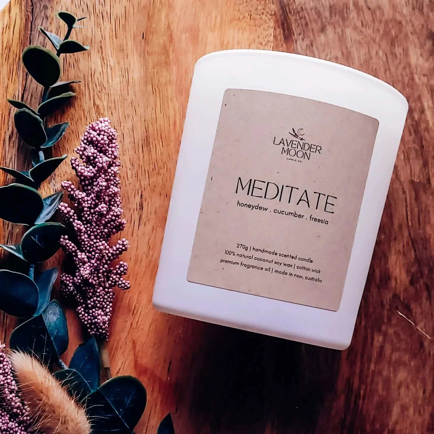 Meditate Candle with honeydew, cucumber, and freesia scent, featuring Rainbow Moonstone chips, vegan coconut soy wax, on a wooden surface with flowers.