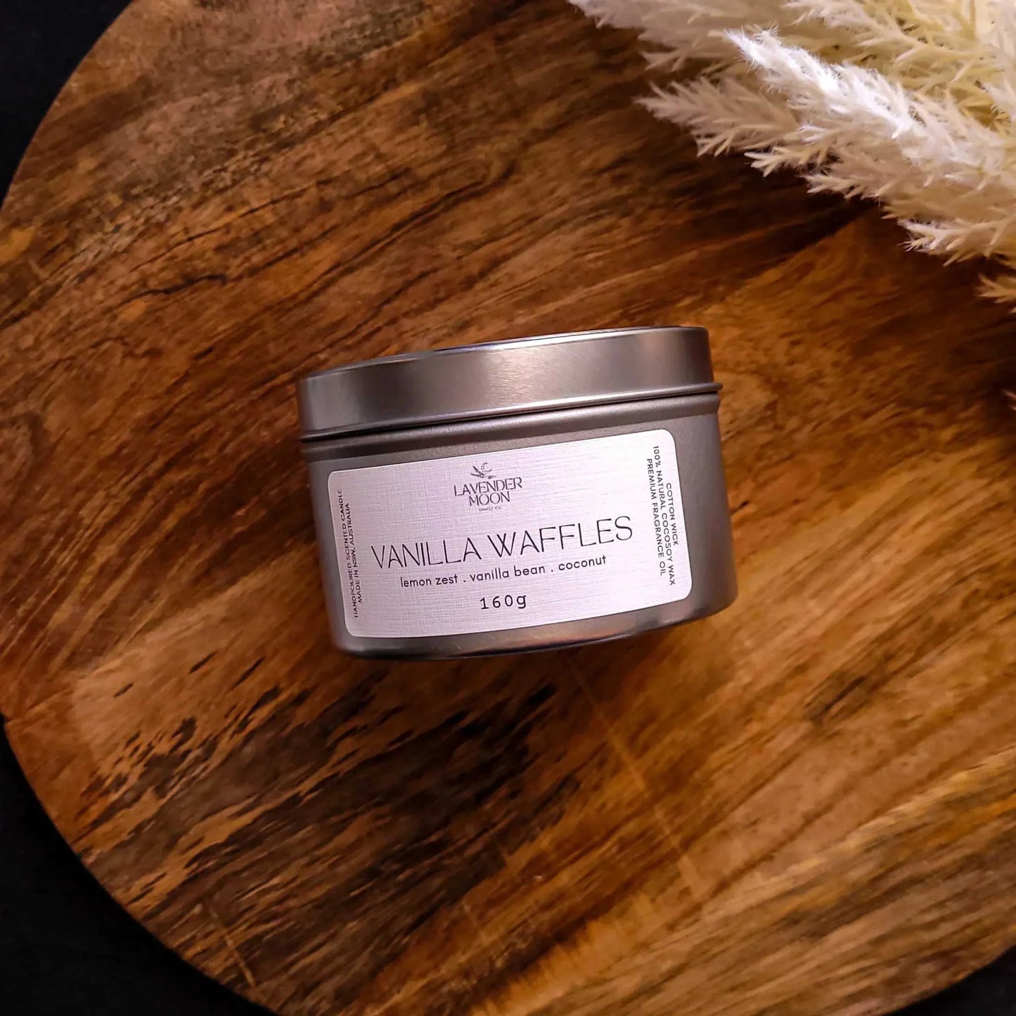 Vanilla Waffles Candle on wooden surface, featuring a tin container with a label indicating lemon zest, vanilla bean, and coconut scents.