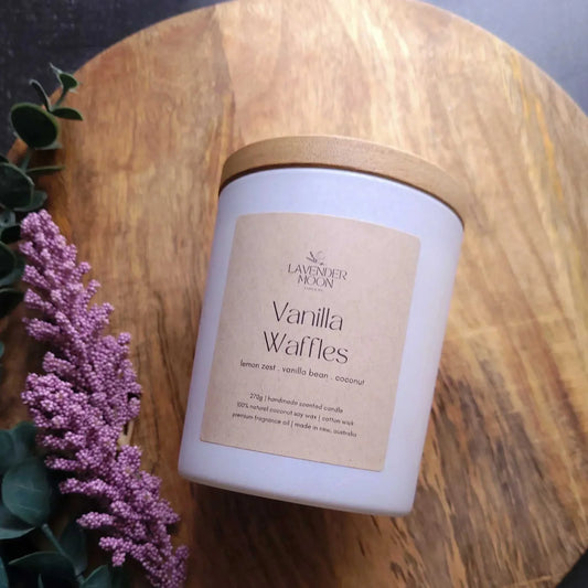 Vanilla Waffles Candle on a wooden surface with purple flowers and greenery.