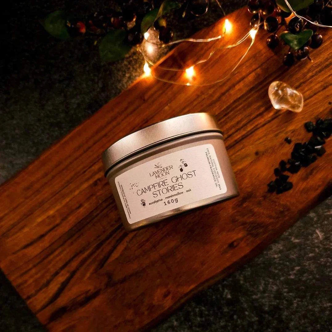 Campfire Ghost Stories 160g candle tin with eucalyptus, marshmallow, and oak scent on a wooden surface adorned with string lights.